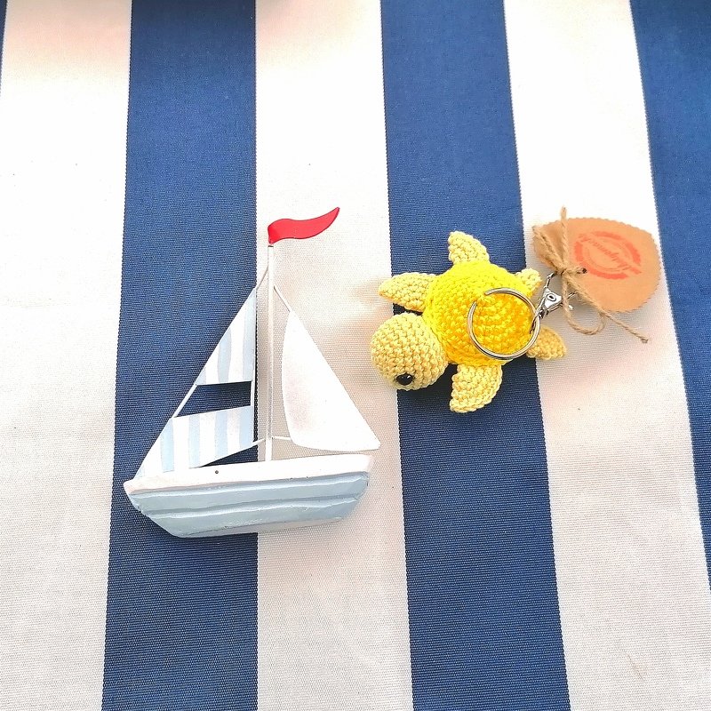 Small yellow crocheted turtle keychain with metal ring beside a wooden toy sailboat on blue-and-white striped fabric