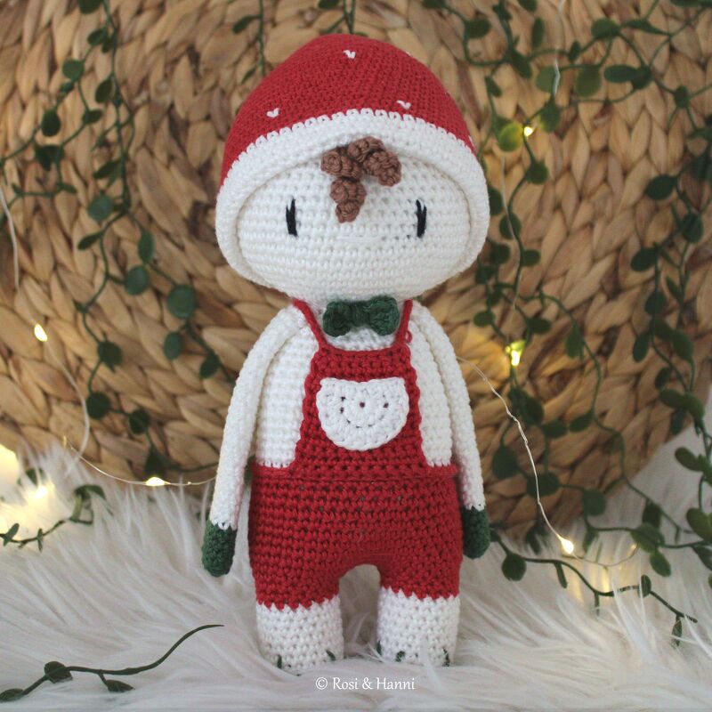 Gehäkelte Glücksbringer-Puppe mit roter Mütze, Latzhose und grüner Fliege vor Lichterkette