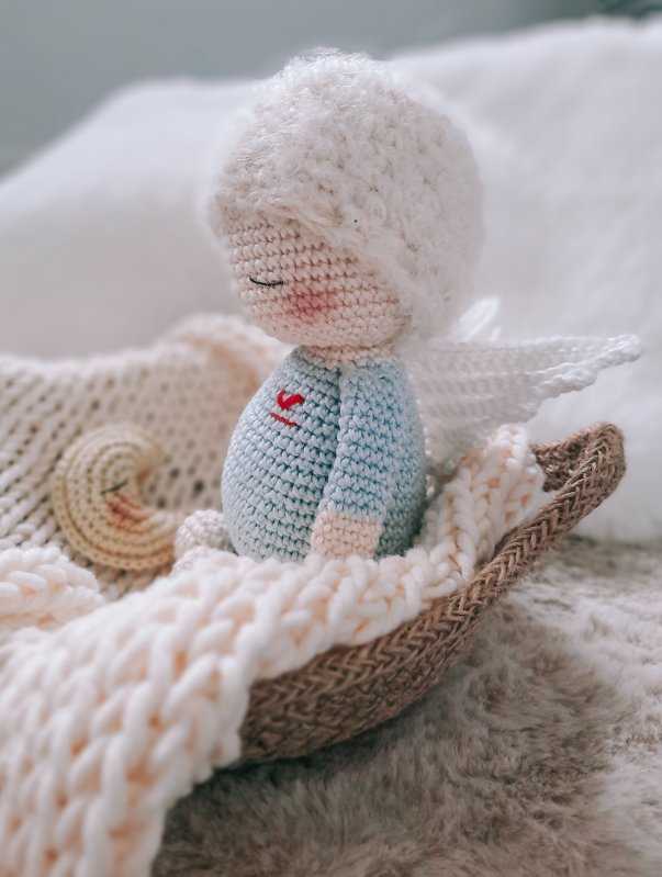 Gehäkeltes Amigurumi-Engelchen mit weißen Flügeln und rotem Herz im Korb auf cremefarbener Decke, mit kleinem Mond
