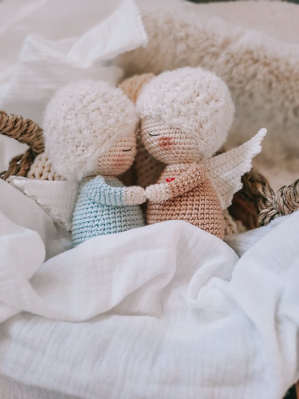 Zwei gehäkelte Amigurumi-Engel mit flauschigem Haar und weißen Flügeln im Korb, sie halten Händchen