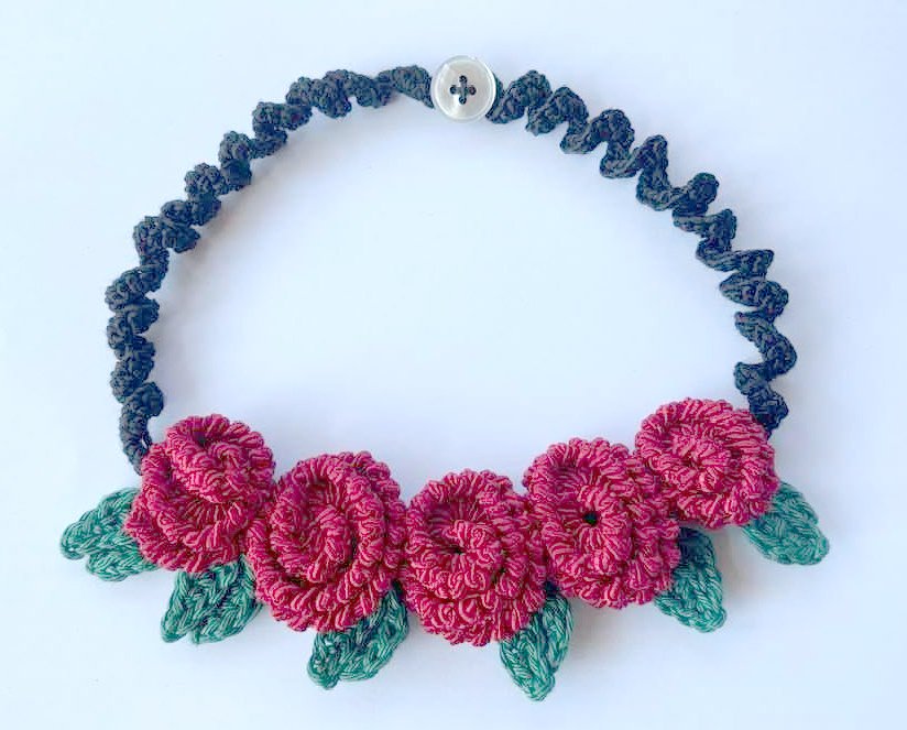 Handgehäkelte Halskette mit fünf roten Blüten, grünen Blättern und dunkelgrauem gedrehtem Band mit Knopfverschluss auf hellem Hintergrund.