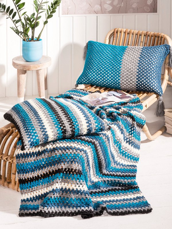 Handgehäkelte Decke mit Zickzack-Streifen in Türkis, Blau, Grau, Schwarz und Weiß mit zwei passenden Kissen auf einem hellen Rattan-Sessel; daneben ein Holzhocker mit Topfpflanze.