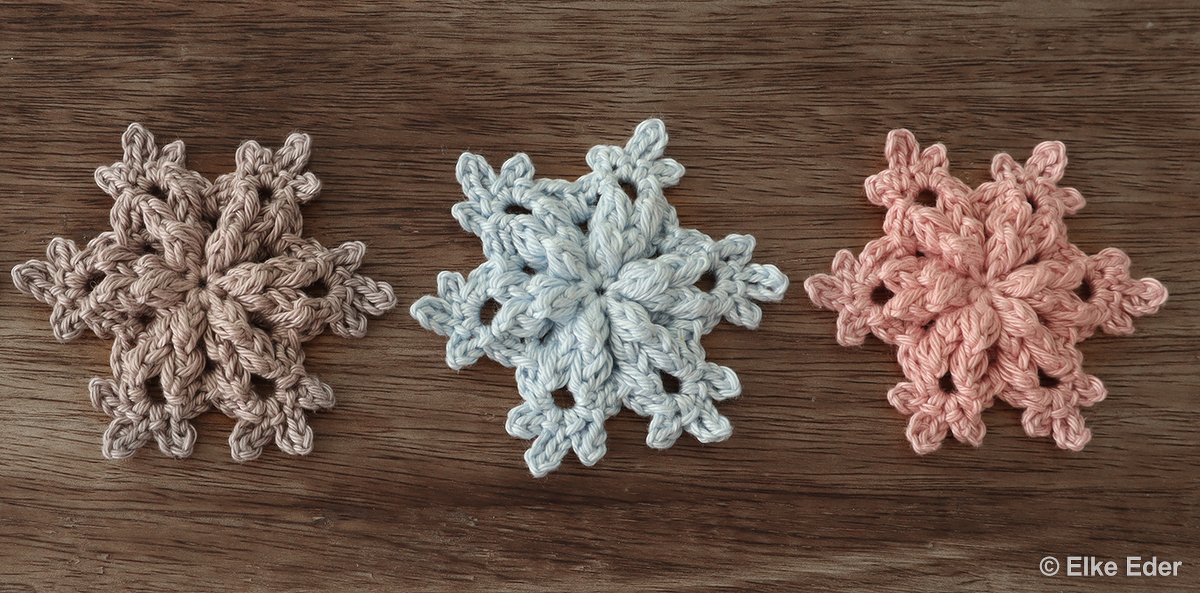 Three crocheted snowflake motifs in beige, light blue and pink on a wooden background