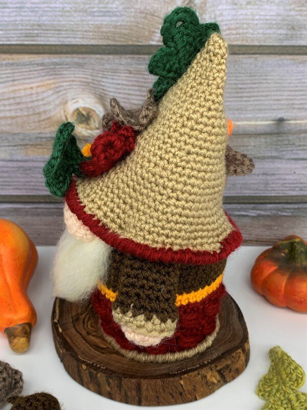 Handcrafted amigurumi gnome with a tall beige hat, autumn leaf accents and white beard on a wooden stand.