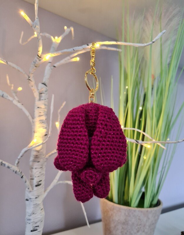 Dunkelrosa gehäkelter Amigurumi-Hase als Schlüsselanhänger an goldfarbenem Karabiner, hängend an einem beleuchteten weißen Zweig.
