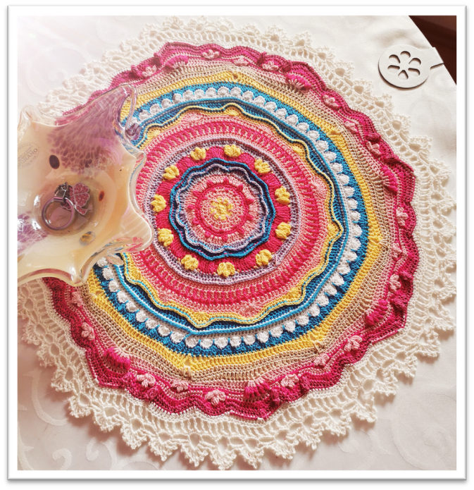 Häkelmandala in Pink, Gelb und Blau mit cremefarbener Spitzenkante auf Tischdecke