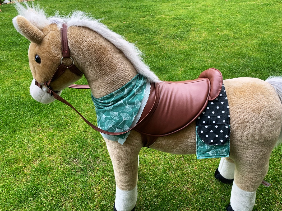 Nahaufnahme: brauner Sattelüberzug mit grüner Halstuchkante und schwarzer, gepunkteter Satteltasche an Plüschpony