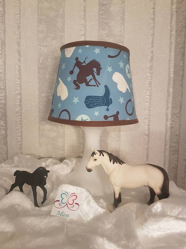Weiße Tischlampe mit blauem Stofflampenschirm mit Pferde-, Hufeisen- und Stiefelmotiven, davor zwei Pferdefiguren