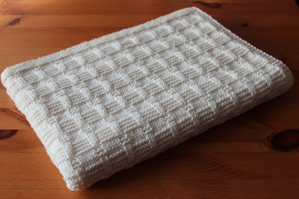 Gestrickte cremeweiße Babydecke mit Waffelstruktur, gefaltet auf einem Holztisch.