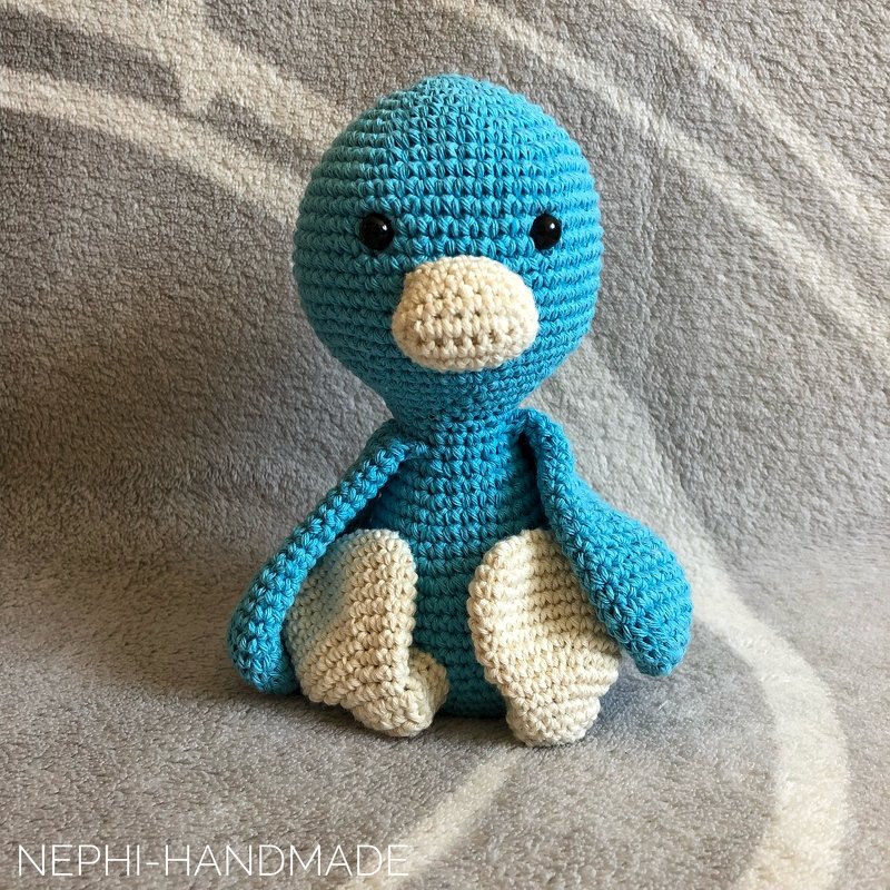 Sitzendes blaues Häkelvogel-Amigurumi mit cremefarbenem Schnabel und Füßen vor neutralem Hintergrund