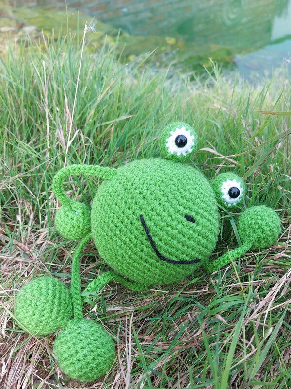 Gehäkelter grüner Amigurumi-Frosch mit großen Augen im Gras am Wasser
