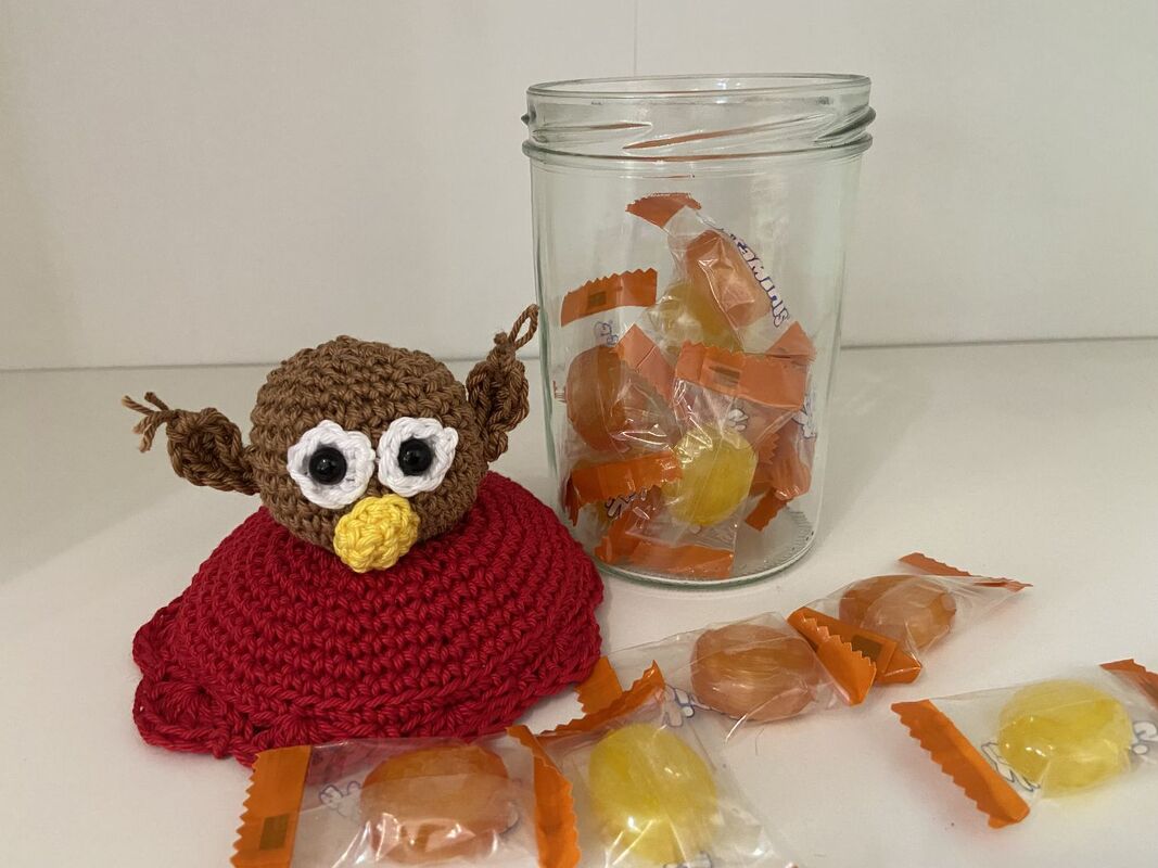 Gehäkelte Gläserhaube in Rot mit braunem Eulenkopf, daneben ein Glas mit verpackten Bonbons