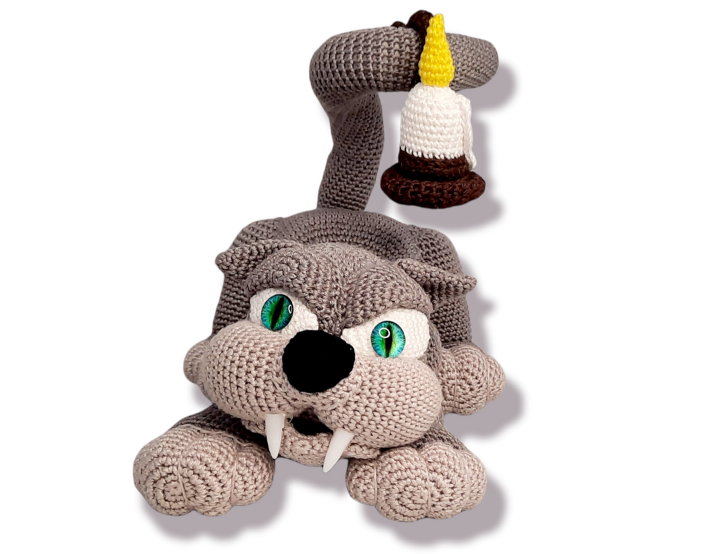 Handmade crocheted saber-toothed cat amigurumi with green eyes, white fangs, and a curled tail holding a small crocheted candle.