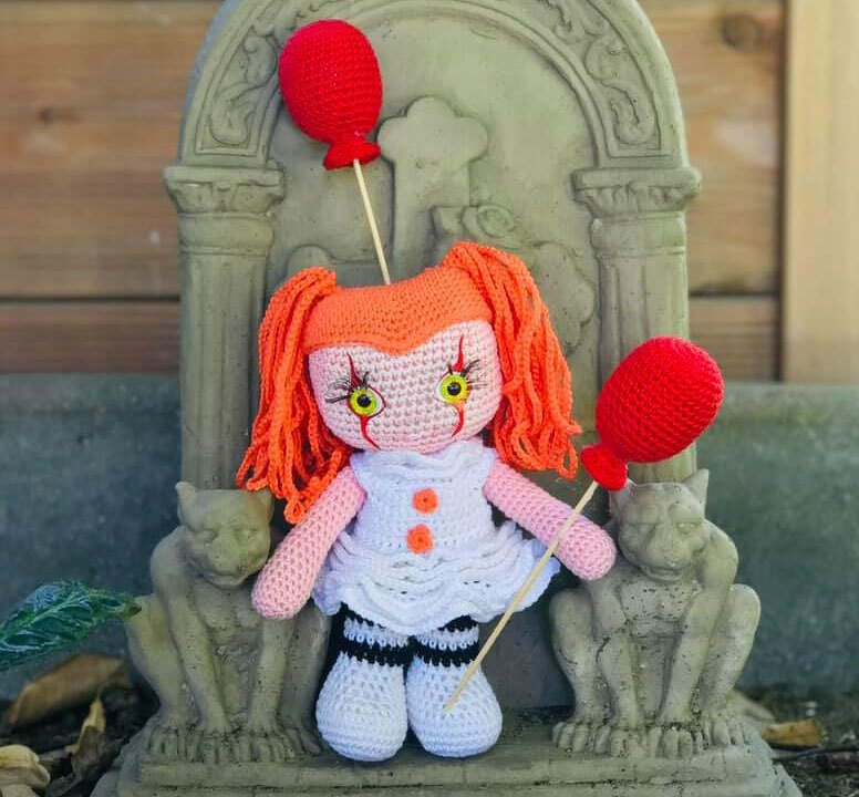 Gehäkelte Clownpuppe mit orangefarbenem Haar und zwei roten Ballons vor einer steinernen Skulptur.