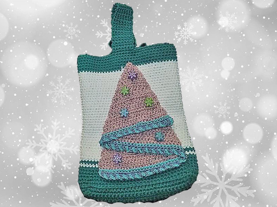 Handgehäkelte Tasche in Grün und Weiß mit appliziertem rosafarbenem Weihnachtsbaum und türkisfarbenen Bändern vor verschneiftem Hintergrund.