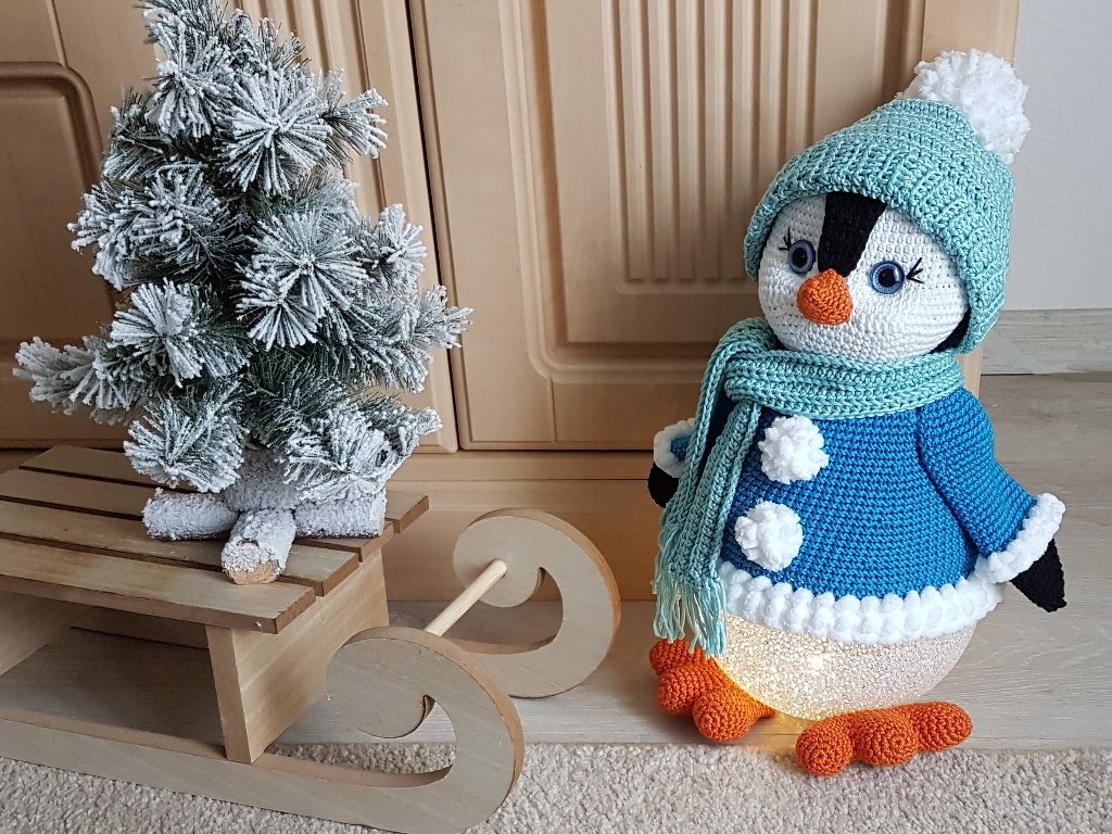 Beleuchteter gehäkelter Pinguin in blauer Jacke und mintgrüner Mütze neben verschneitem Mini-Weihnachtsbaum auf Holzschlitten