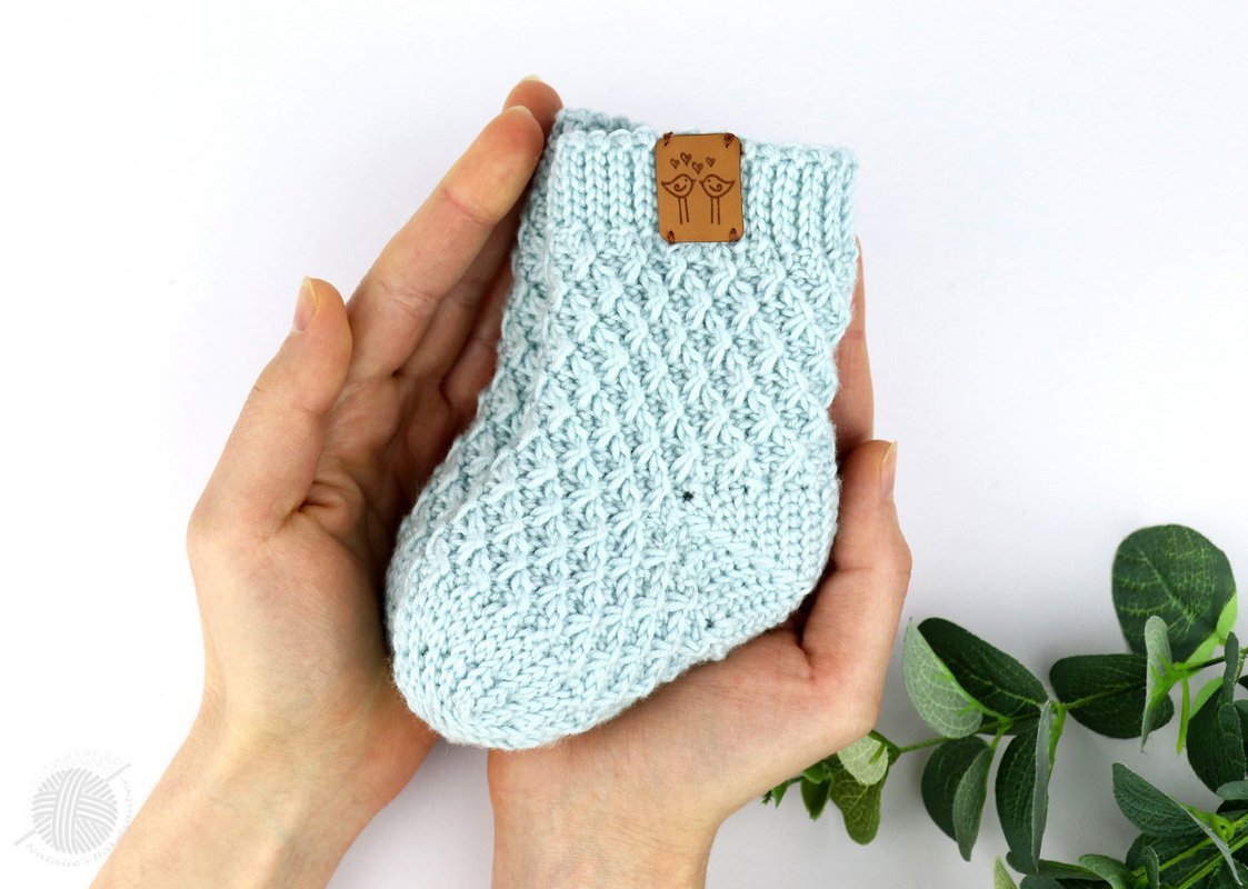 Hands holding a light blue crocheted baby sock with a textured stitch pattern and a small brown bird tag on the ribbed cuff.