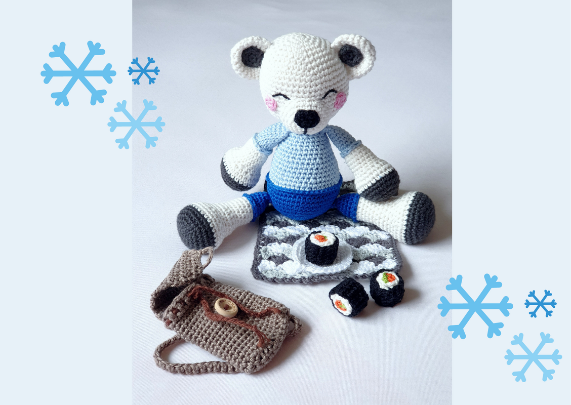 Gehäkelter weißer Eisbär in blauem Outfit mit drei Mini-Sushi und brauner gehäkelter Tasche auf einer Matte