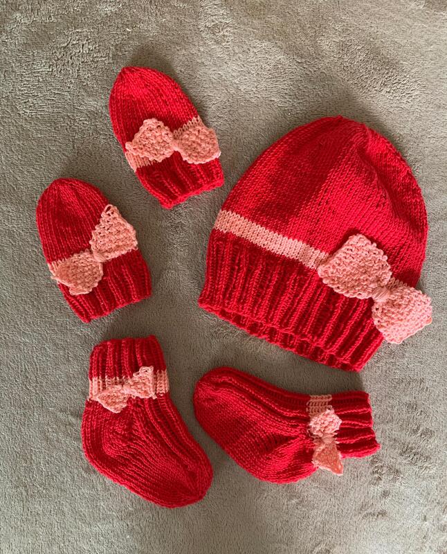 Rotes und rosa gestricktes Baby-Set mit Schleifen: eine große Mütze, zwei kleine Mützen und ein Paar Socken auf grauem Untergrund