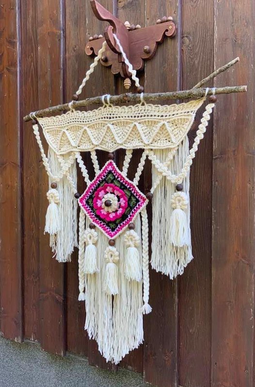 Cremefarbener gehäkelter Wandbehang mit Quasten und pink-grünem Rautenmuster, an einem Ast vor Holzwand