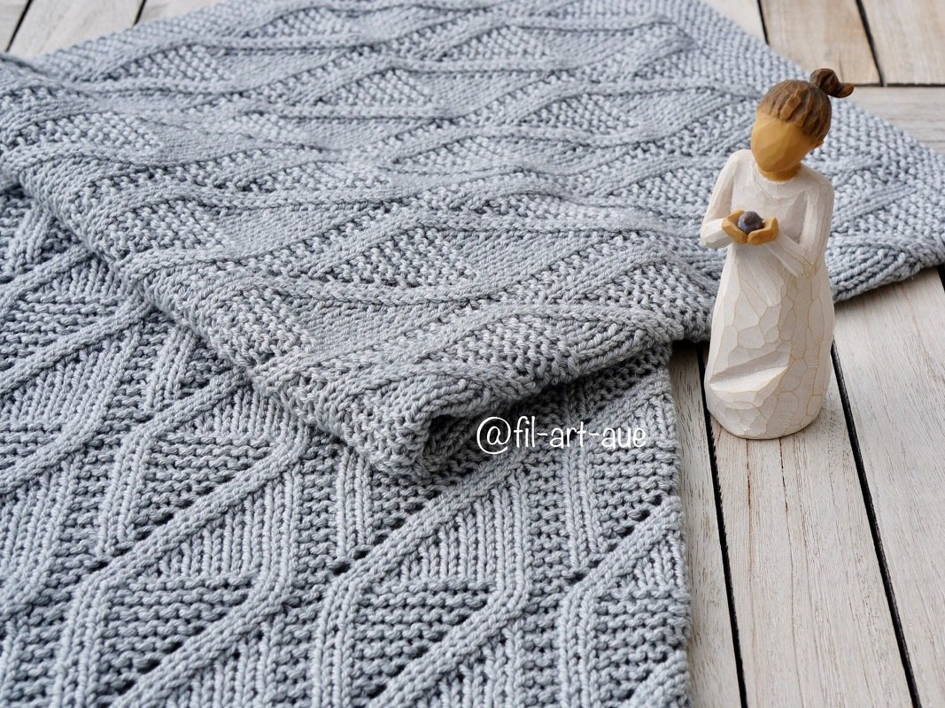 Light gray knitted baby blanket with diamond stitch pattern folded on wooden boards with a small carved wooden figure