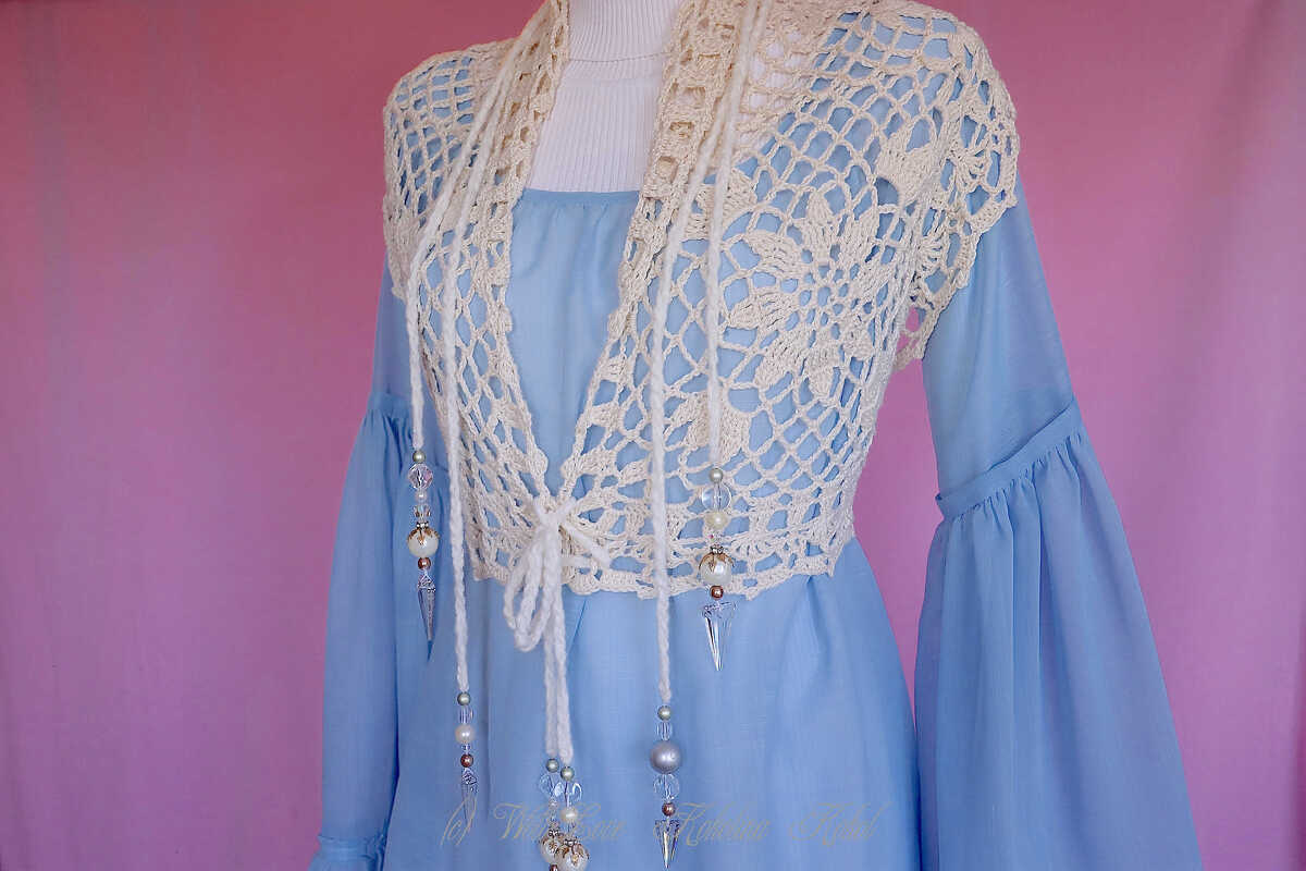 Cremefarbene gehäkelte Weste mit Lochmuster und perlenbesetzten Bändern über einem hellblauen Kleid auf einer Schaufensterpuppe.