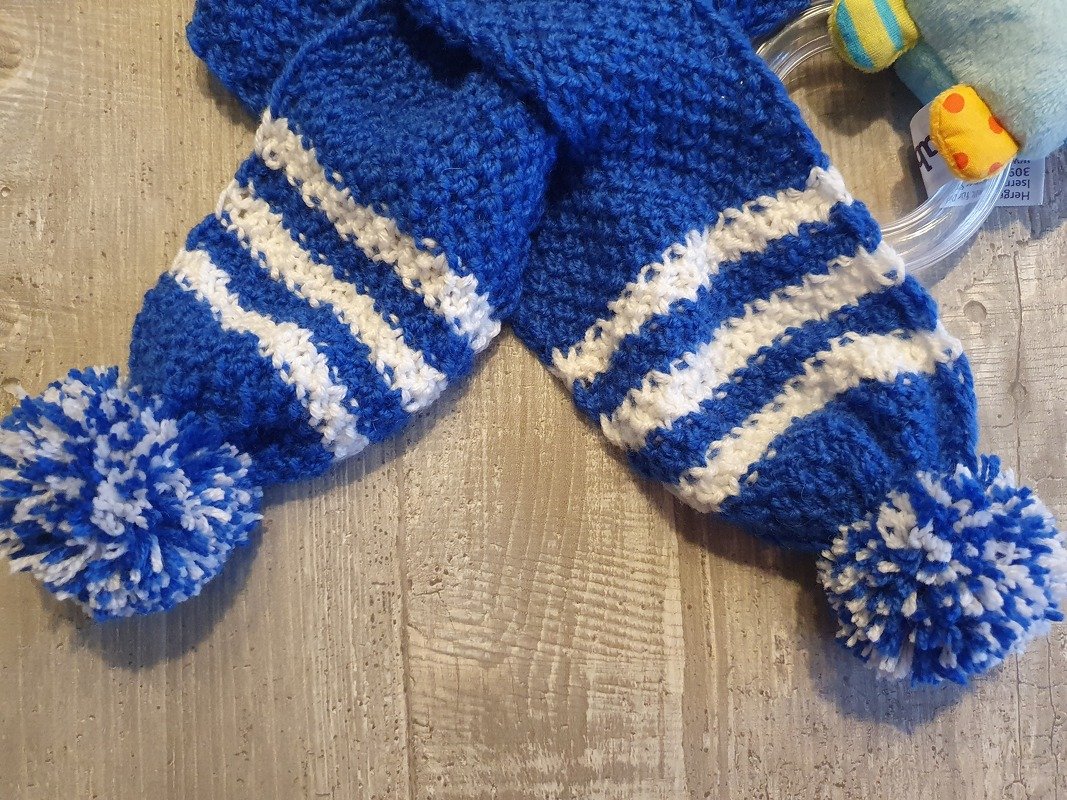 Gestrickter blau-weißer Babyschal mit zwei Bommeln auf Holzuntergrund