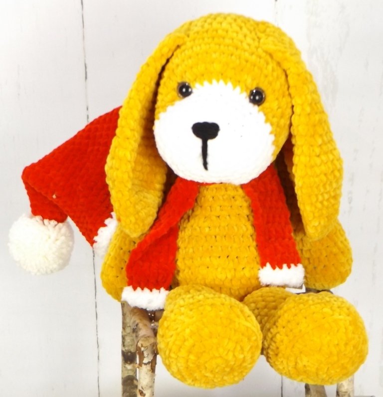 Gehäkelter gelber Amigurumi-Hund mit langen Schlappohren, weißer Schnauze, schwarzen Augen und Nase, roter Weihnachtsmütze mit Pompon und rotem Schal vor hellem Hintergrund.