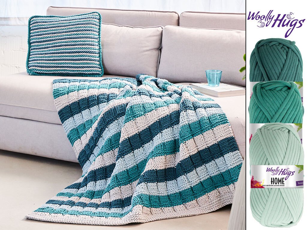 Gestrickte Decke und quadratisches Kissen in Türkis-, Blau- und Weißstreifen auf hellem Sofa; drei Garnknäuel daneben.