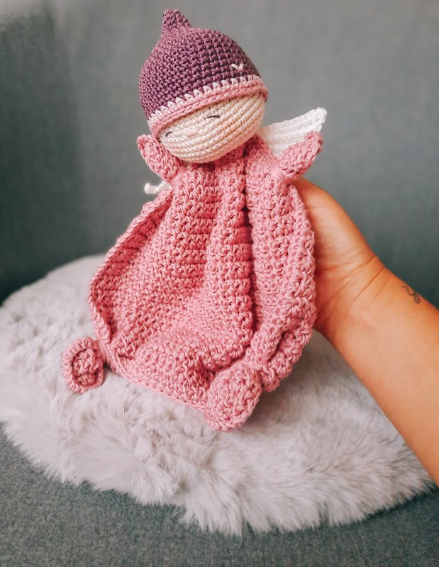 Hand holding pink crocheted baby lovey with small doll head wearing a purple cap on a faux fur surface