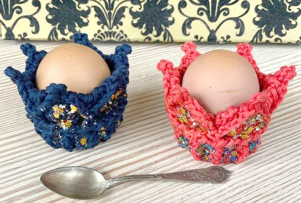 Zwei gehäkelte Eierwärmer in Blau und Koralle mit Glitzerdetails, jeweils ein braunes Ei; ein Silberlöffel liegt auf einem hellen Holztisch.