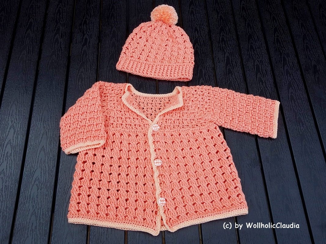 Gehäkelte Babyjacke mit Knöpfen und Korallen‑Bommelmütze auf dunklem Holz