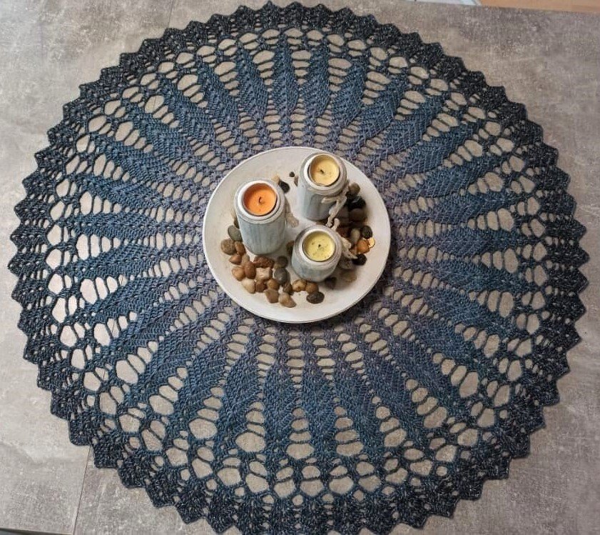 Rundes gehäkeltes Deckchen in Blau- und Schwarztönen mit Spitzenmuster und gezacktem Rand. In der Mitte ein Teller mit drei Kerzen und Kieselsteinen.