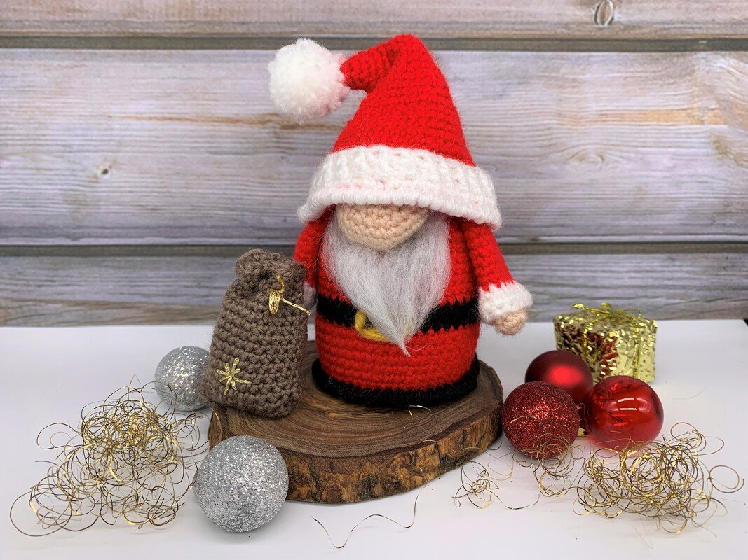 crocheted Santa amigurumi with red hat and white beard on wood slice next to a small brown sack and red and silver baubles