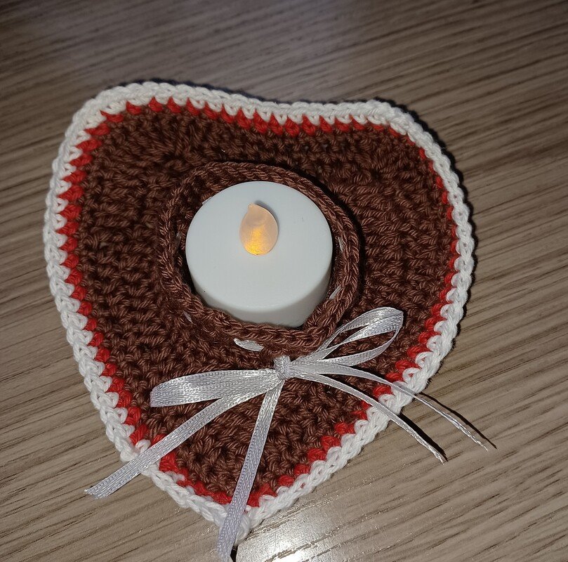 Herzförmiger gehäkelter Untersetzer in Braun mit roten und weißen Rändern, LED-Teelicht in der Mitte und silberner Schleife