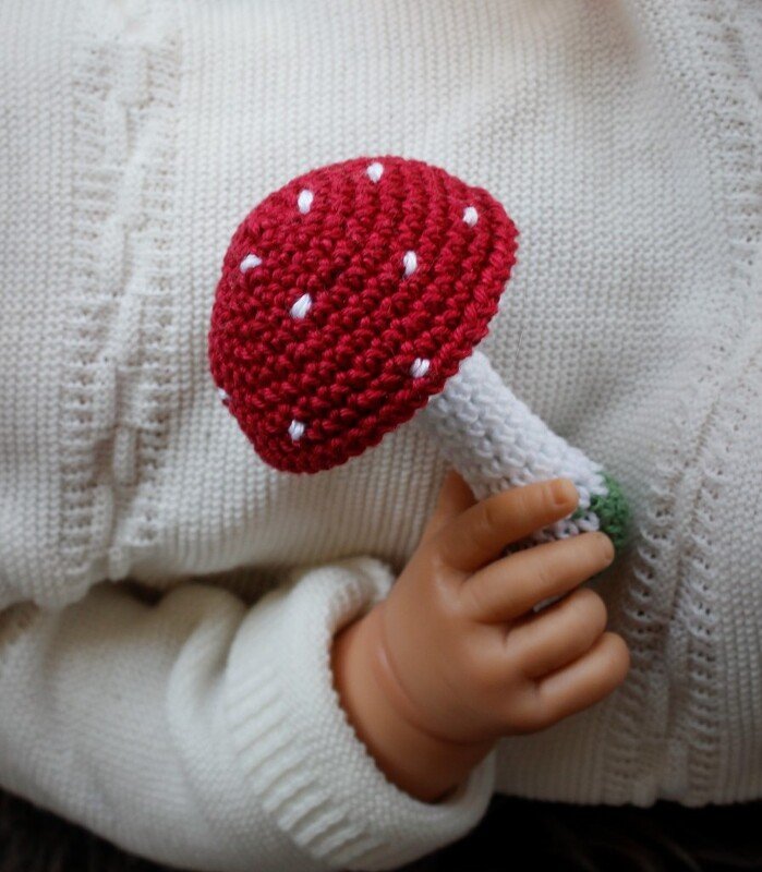 Handgehäkelter roter Pilz mit weißen Punkten, gehalten von einer Babyhand vor weißem Strick.