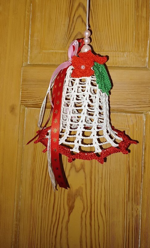 Gehäkelte weiße Glocke mit roter Kante, Blüte, grünem Blatt und Bändern, hängend an Holztür