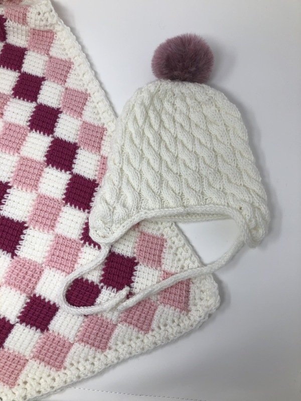 Nahaufnahme einer weißen gestrickten Ohrenklappenmütze mit Zopfmuster, Bindebändern und rosa Bommel auf karierter Decke