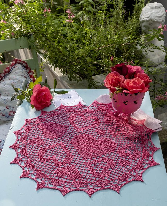 Rosa rundes gehäkeltes Deckchen mit Herzmuster auf hellblauem Tisch, dekoriert mit Rosen.