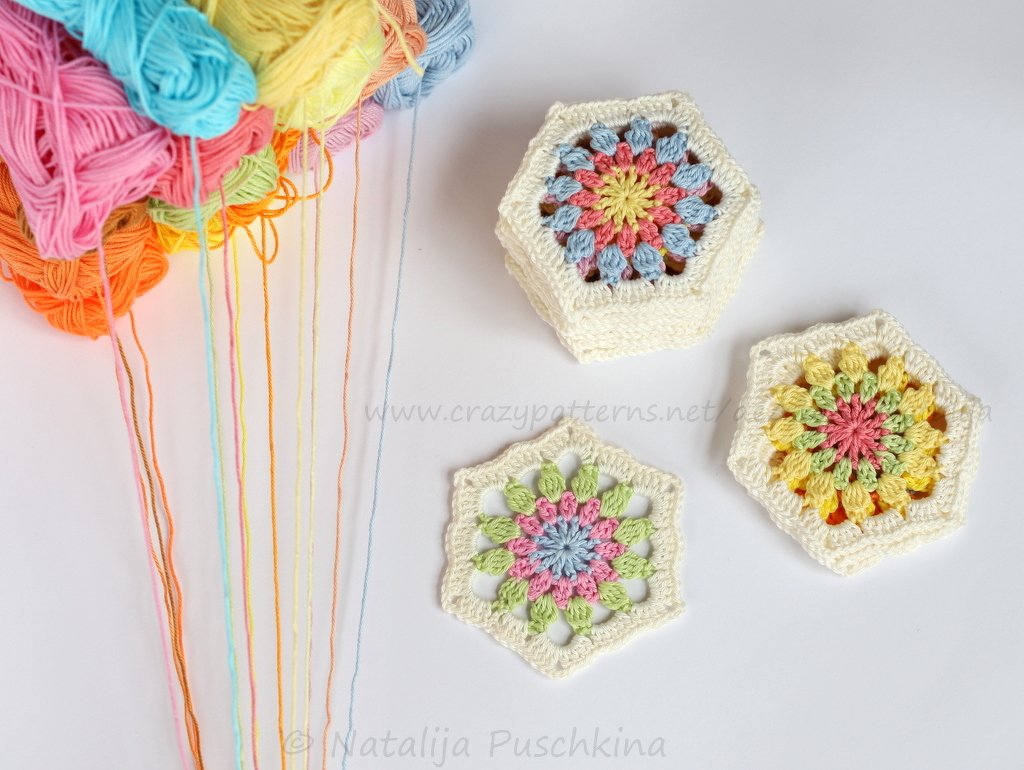 Drei gehäkelte Hexagon-Grannymotive in Pastelltönen neben Garnknäueln mit herabhängenden Fäden