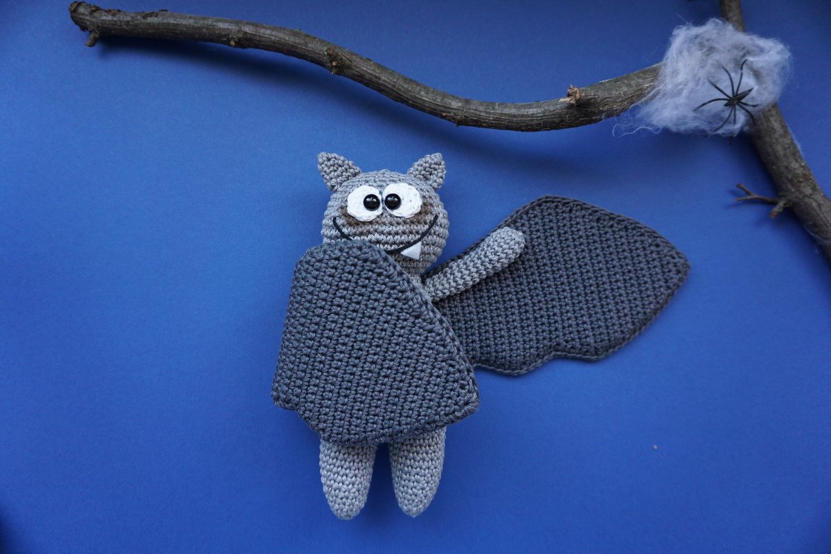 Niedliche graue Fledermaus-Amigurumi mit Flügeln neben Ast und Spinnennetzdekoration