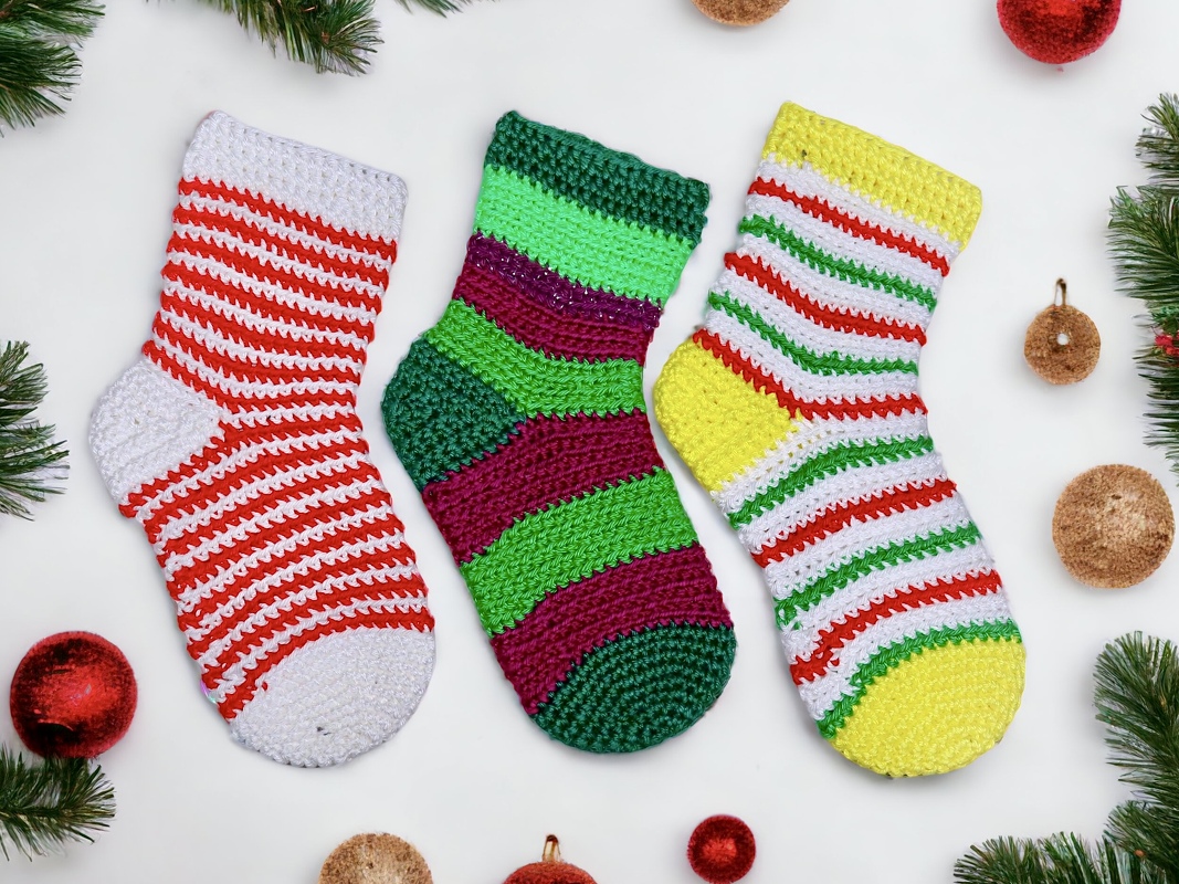 Three striped crochet Christmas stockings with varied colorways, placed beside pine sprigs and round ornaments.