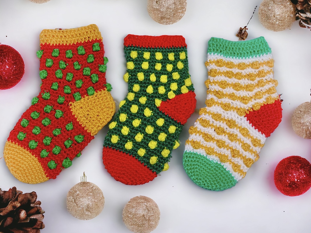 Drei kleine gehäkelte Weihnachtsstrümpfe in Rot, Grün und Gelb mit Noppenmuster, umgeben von Kugeln und Tannenzapfen.