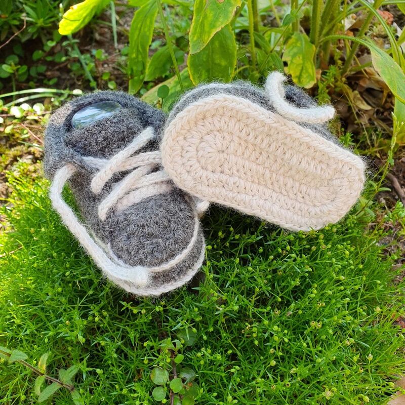 Zwei gehäkelte Baby-Sneaker in Grau und Weiß mit Schnürung auf grünem Gras, ein Schuh mit sichtbarer heller Sohle