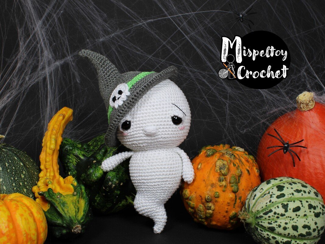 White crocheted ghost amigurumi wearing a grey witch hat with skull accent, shown with pumpkins, gourds and decorative spiderweb backdrop.