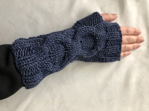 Hand mit blau gestrickter, fingerloser Armstulpe mit Zopfmuster und Daumenloch auf hellem Hintergrund