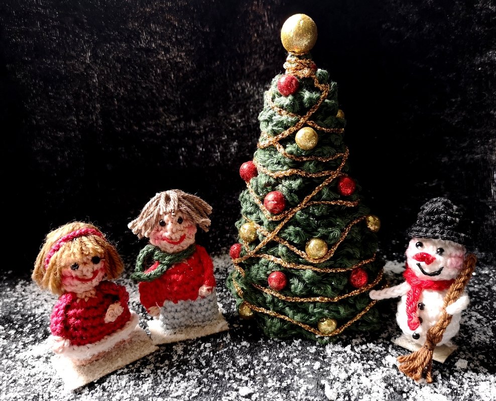 Mini-Weihnachtsbaum aus grünem Garn mit goldener Girlande, zwei gehäkelte Kinderfiguren und ein Schneemann auf schwarzer Fläche mit Kunstschnee.