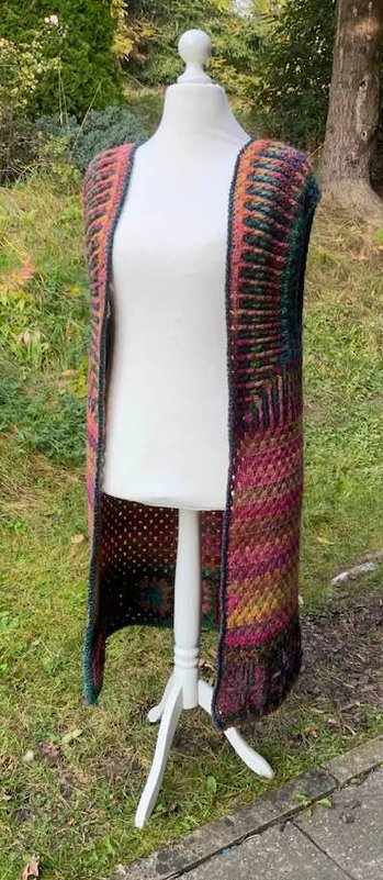 Lange, offene gehäkelte Damenweste in bunten Farbverläufen auf weißer Schneiderpuppe vor grünem Hintergrund.