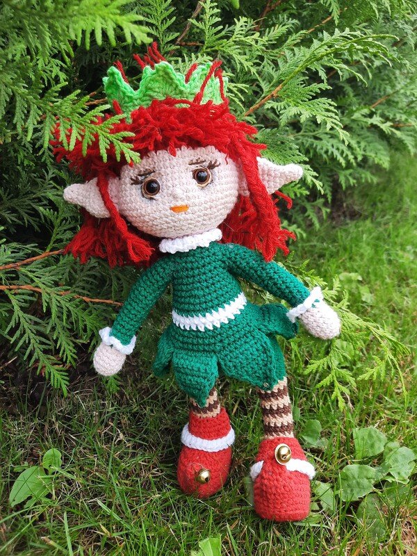 Verspielte Amigurumi-Waldelfe mit Blattkrone, rotem Haar und grünem Kleidchen vor einem Nadelbusch