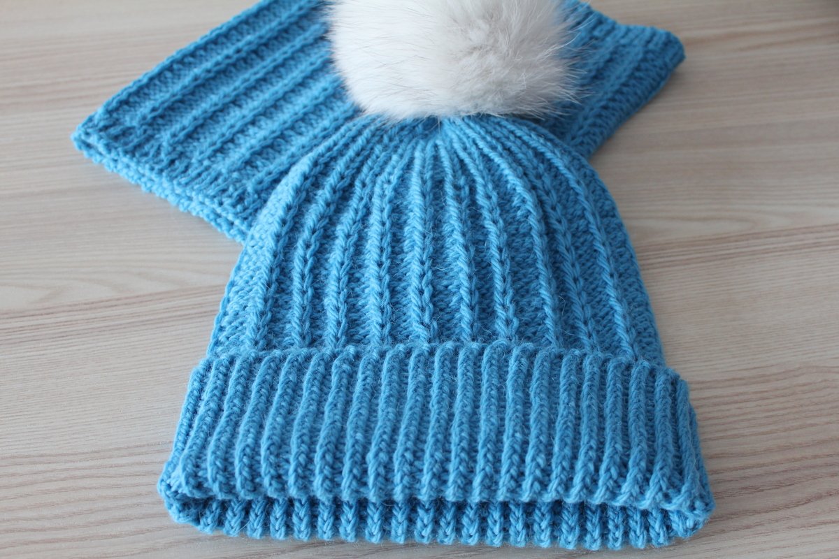 Blaues, geripptes Mützen- und Schalset mit weißem Bommel auf hellem Holz